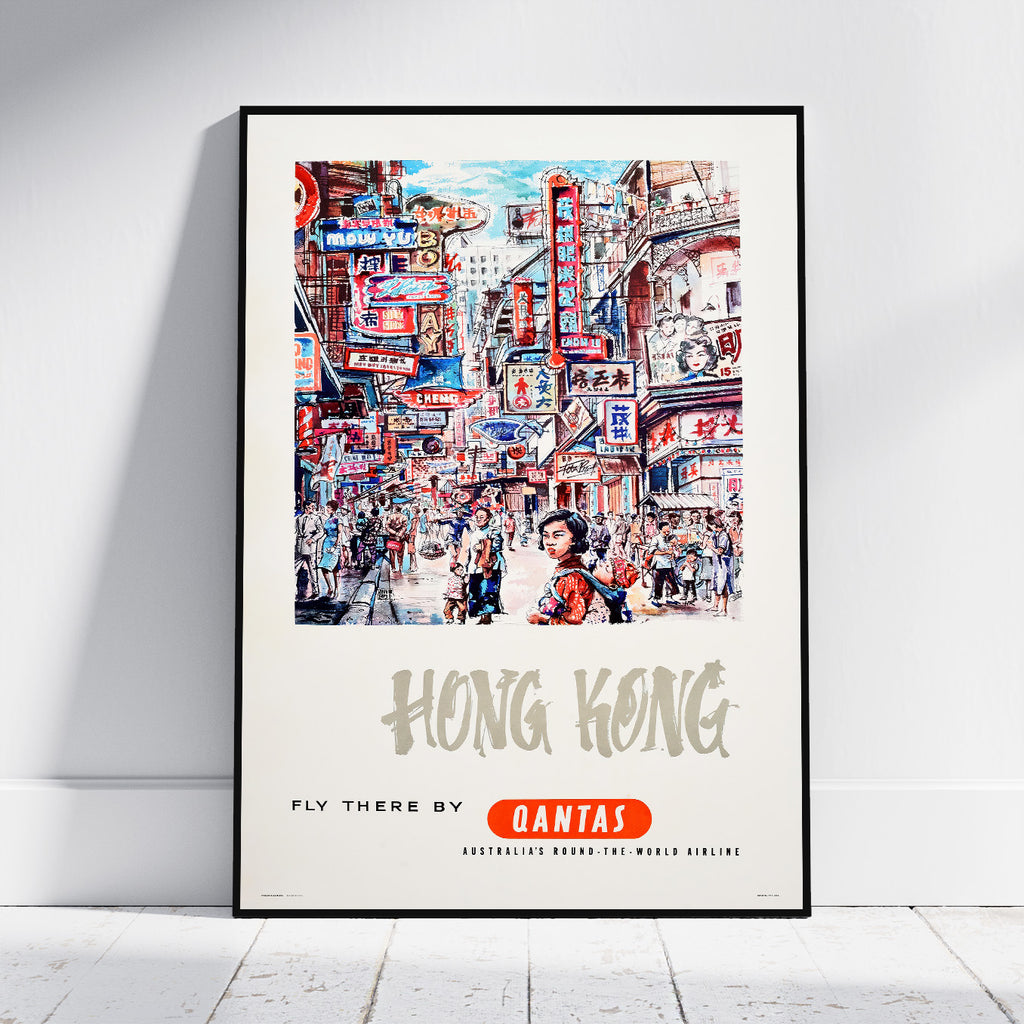 A framed vintage-style travel poster of Hong Kong, leaning against a white wall on a light wood floor. The poster features a colorful illustration of a busy street scene with the text 'HONG KONG' and 'FLY THERE BY QANTAS'.