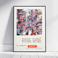 A framed vintage-style travel poster of Hong Kong, leaning against a white wall on a light wood floor. The poster features a colorful illustration of a busy street scene with the text 'HONG KONG' and 'FLY THERE BY QANTAS'.