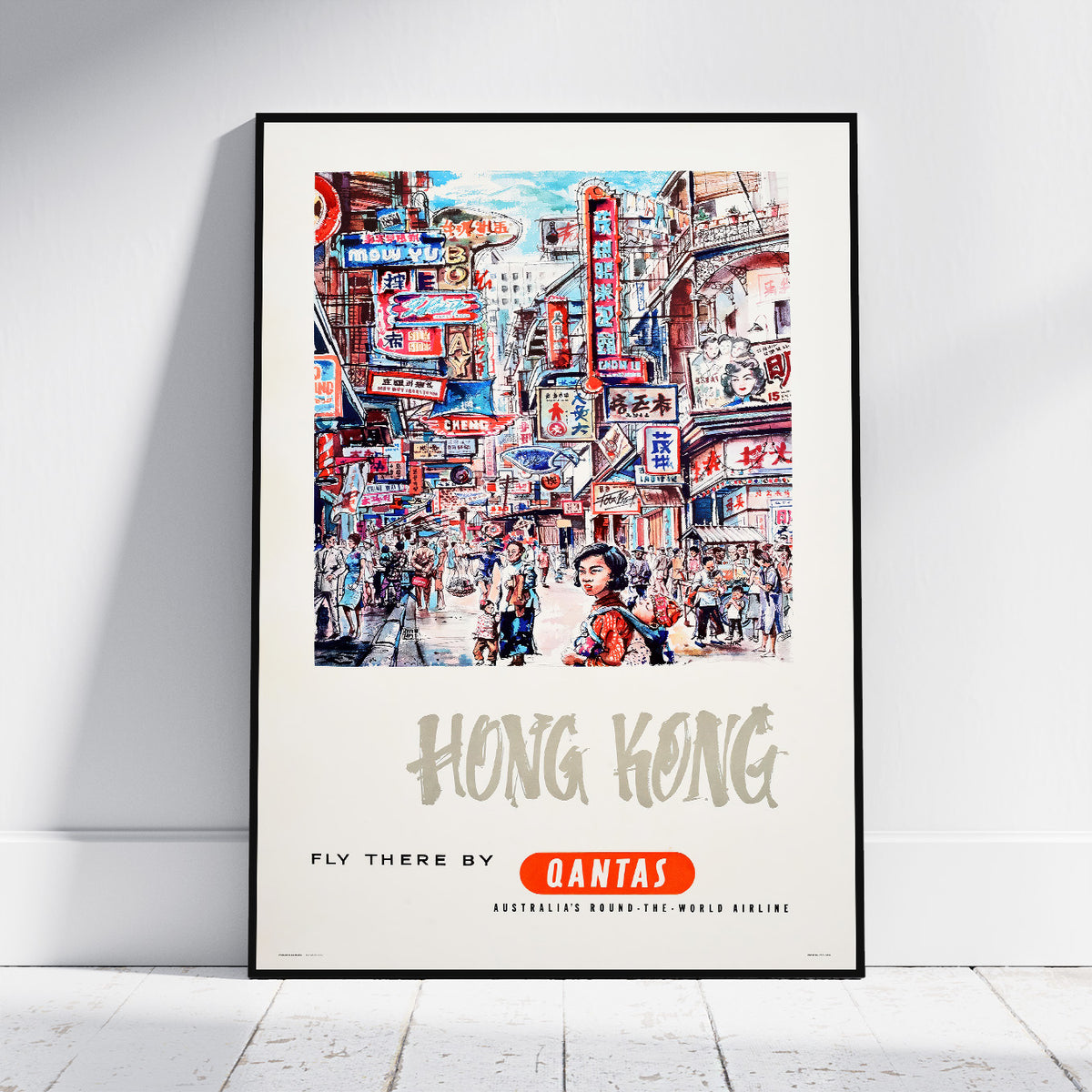A framed vintage-style travel poster of Hong Kong, leaning against a white wall on a light wood floor. The poster features a colorful illustration of a busy street scene with the text 'HONG KONG' and 'FLY THERE BY QANTAS'.