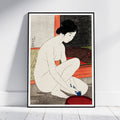 A framed art print of a Japanese ukiyo-e woodblock illustration of a nude woman washing her feet, leaning against a white wall on a light wood floor.