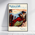 A framed vintage-style poster leaning against a white wall on a light floor. The poster features the word 'Venise' at the top and an illustrated scene with a woman in a red cloak and a man in a mask near water and a lighthouse.