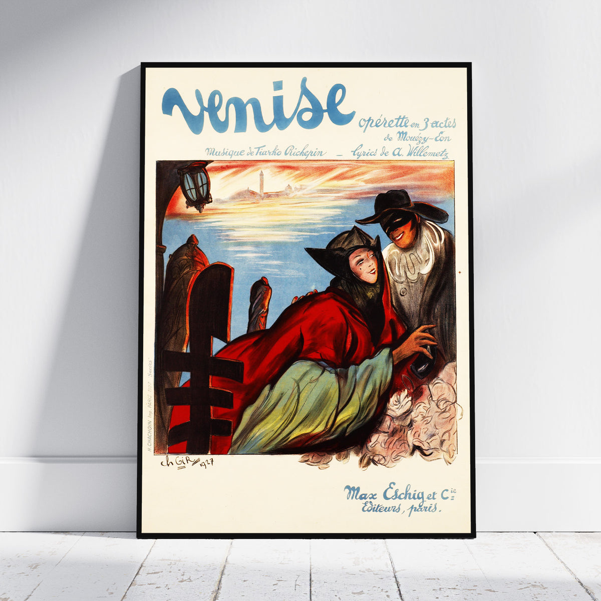 A framed vintage-style poster leaning against a white wall on a light floor. The poster features the word 'Venise' at the top and an illustrated scene with a woman in a red cloak and a man in a mask near water and a lighthouse.