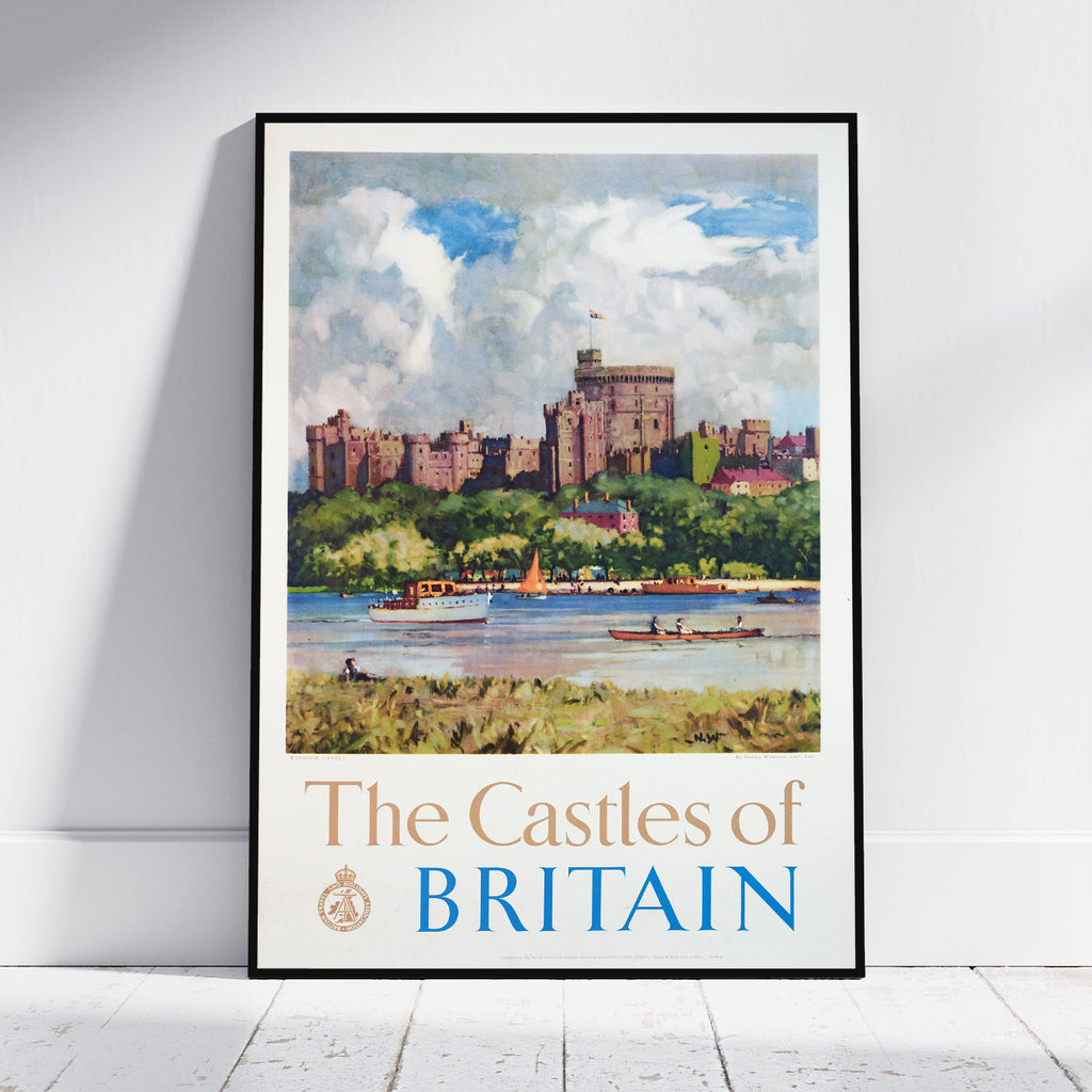 A framed travel poster print of 'The Castles of Britain' leaning against a white wall on a light wood floor. The poster features a watercolor-style illustration of a castle on a hill, boats on the water, and the title 'The Castles of BRITAIN' at the bottom.