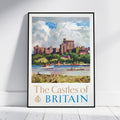 A framed travel poster print of 'The Castles of Britain' leaning against a white wall on a light wood floor. The poster features a watercolor-style illustration of a castle on a hill, boats on the water, and the title 'The Castles of BRITAIN' at the bottom.