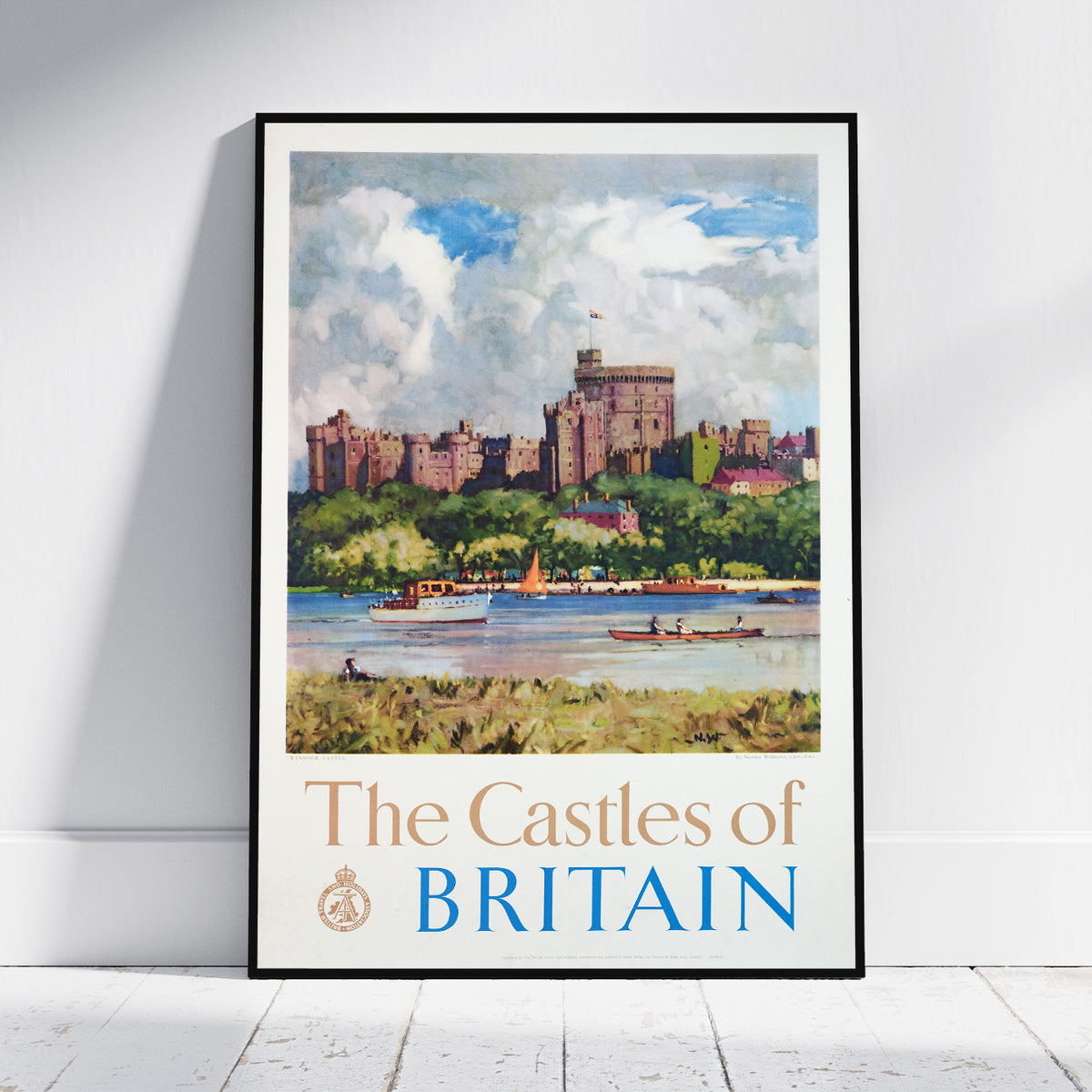 A framed travel poster print of 'The Castles of Britain' leaning against a white wall on a light wood floor. The poster features a watercolor-style illustration of a castle on a hill, boats on the water, and the title 'The Castles of BRITAIN' at the bottom.