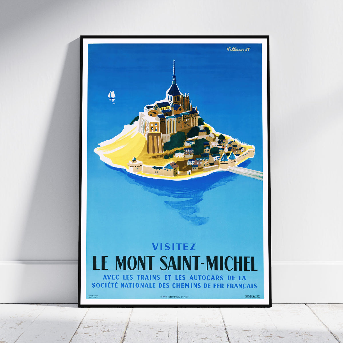 A framed vintage-style travel poster of Le Mont Saint-Michel, France, leaning against a white wall on a light wood floor. The poster features a bright blue background with an illustration of the island and cathedral, plus French text at the bottom.