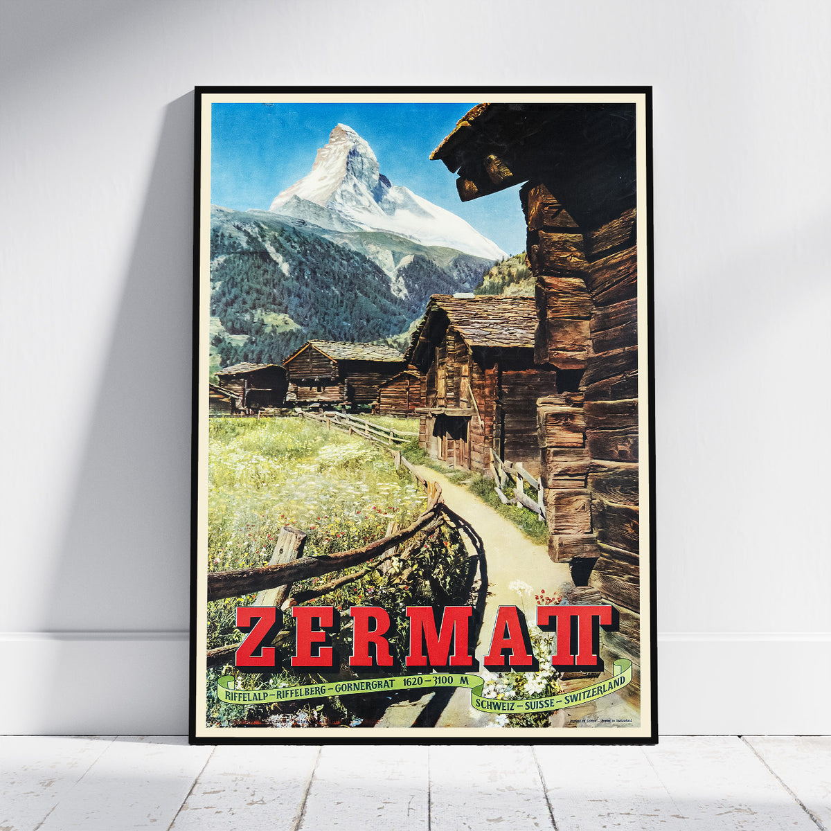 A framed vintage-style travel poster leaning against a white wall on a light floor. The poster depicts a mountain village with rustic wooden cabins, a grassy field, and a snow-capped mountain peak under a blue sky.
