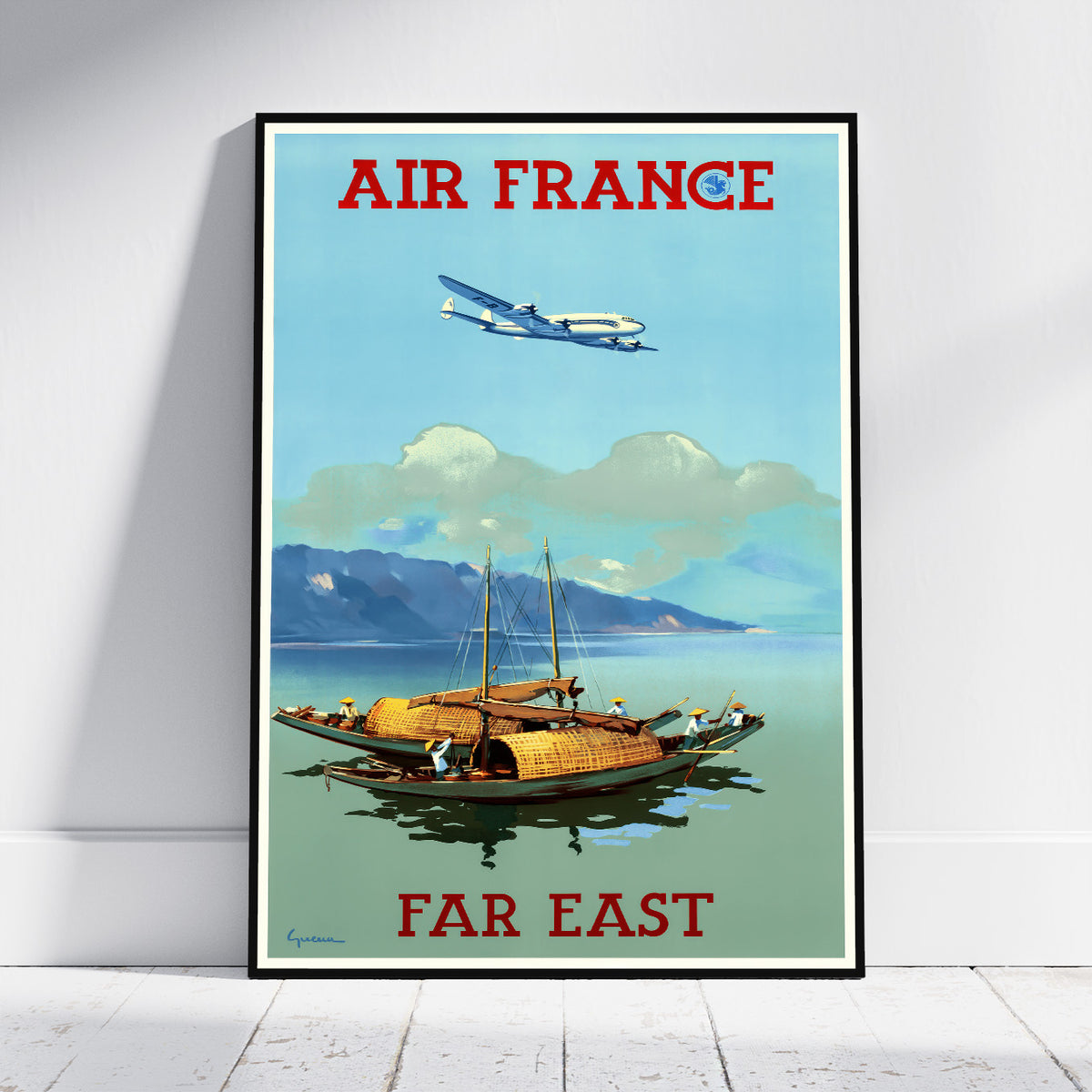 A framed vintage-style Air France travel poster leaning against a white wall on a light wood floor. The poster shows the words 'AIR FRANCE' at the top and 'FAR EAST' at the bottom, with a blue airplane flying above a coastal scene featuring mountains, water, and boats.
