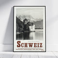 A framed vintage-style travel poster of Switzerland leaning against a white wall on a light wood floor. The poster features a black-and-white photograph of a castle by the water with mountains in the background, and large red 'SCHWEIZ' text at the bottom.