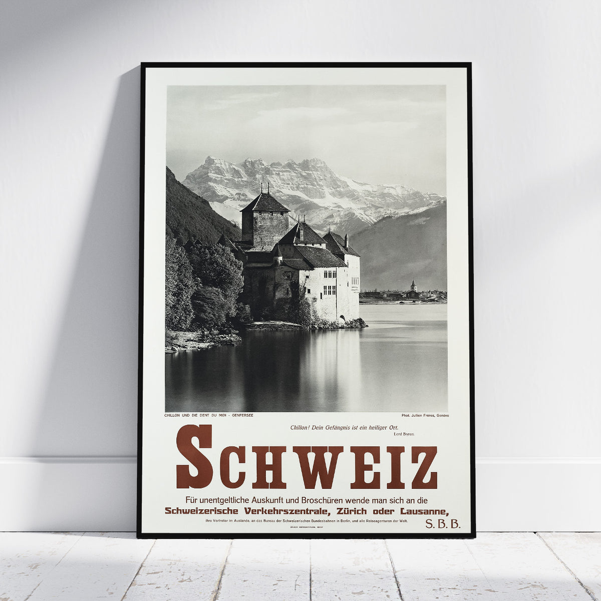A framed vintage-style travel poster of Switzerland leaning against a white wall on a light wood floor. The poster features a black-and-white photograph of a castle by the water with mountains in the background, and large red 'SCHWEIZ' text at the bottom.