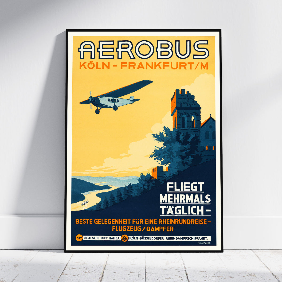 A framed vintage-style travel poster leaning against a white wall on a light wood floor. The poster features an illustration of an airplane flying over a coastal landscape with a tower and buildings, and large text reading 'AEROBUS KÖLN - FRANKFURT/M' and 'FLIEGT MEHRMALS TÄGLICH'.