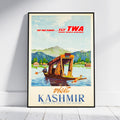 A vintage-style travel poster for TWA Trans World Airlines advertising Kashmir, leaning against a white wall on a light wood floor. The poster features an illustration of a wooden boat on a lake with mountains in the background, with red and blue text.