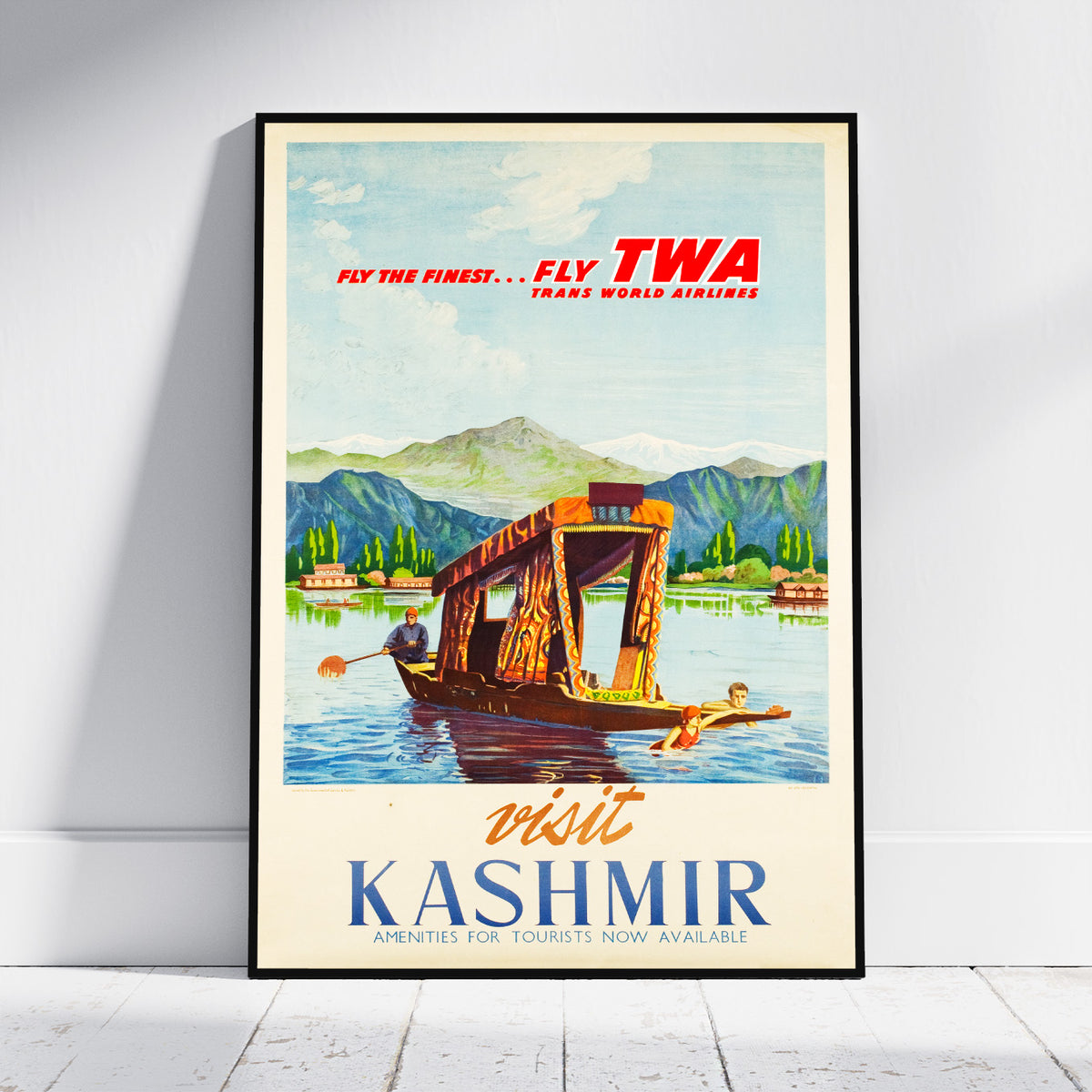 A vintage-style travel poster for TWA Trans World Airlines advertising Kashmir, leaning against a white wall on a light wood floor. The poster features an illustration of a wooden boat on a lake with mountains in the background, with red and blue text.