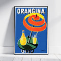 A framed vintage-style poster leaning against a white wall on a light floor. The poster shows the word "ORANGINA" at the top, with a large spiral orange peel under a black umbrella, a yellow-orange bottle, and a glass of orange drink on a blue background.