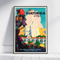 A framed vintage-style travel poster leaning against a white wall on a light floor. The poster shows a woman carrying a red hat in front of a large stepped temple, with the text 'Fly to GUATEMALA by El Chapo' and 'Pan American World Airways'.