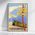 A framed vintage travel poster of Lötschberg, Switzerland, leaning against a white wall on a light wood floor. The poster features a retro-style illustration of a train traveling through the mountains under a blue sky, with a prominent yellow tree in the foreground.