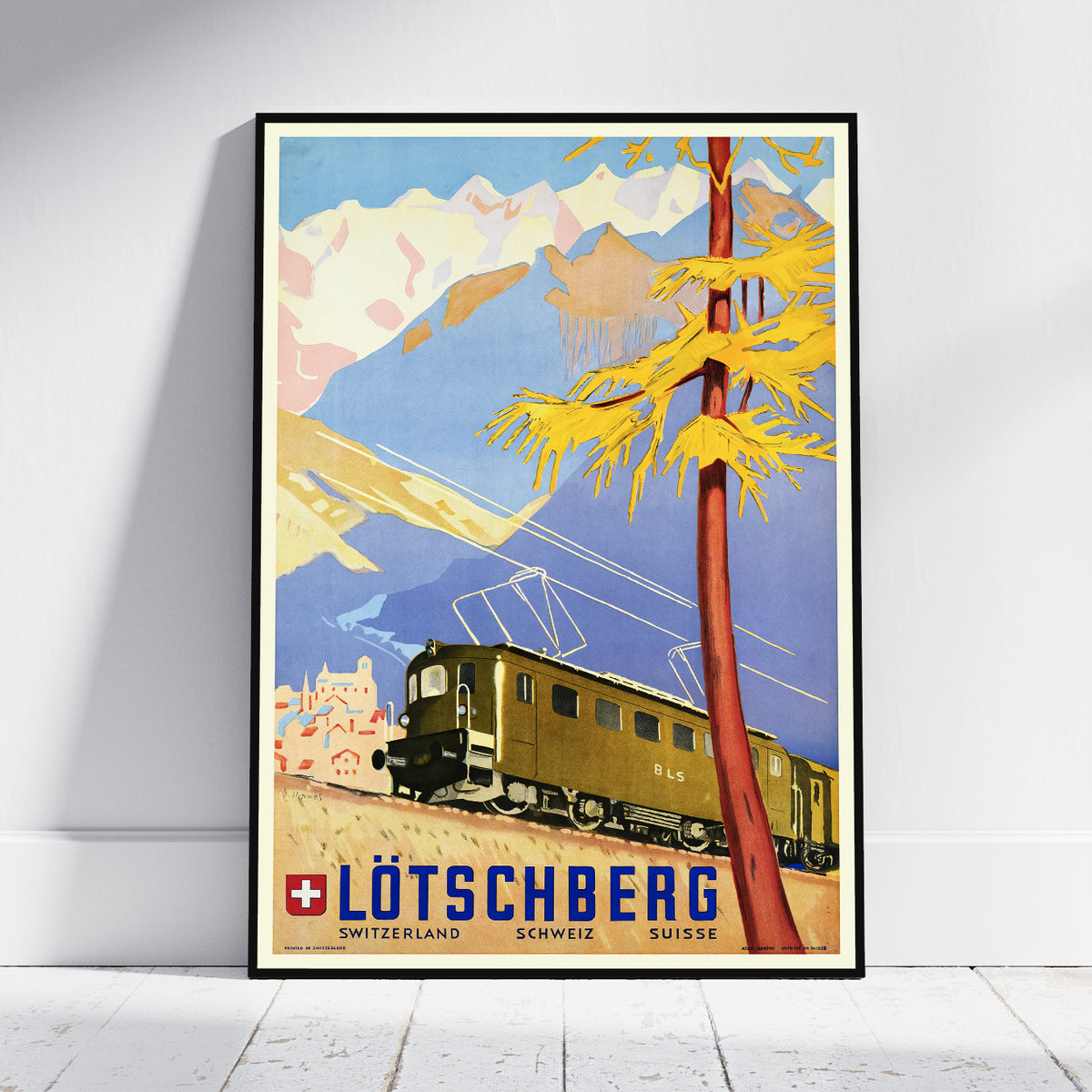 A framed vintage travel poster of Lötschberg, Switzerland, leaning against a white wall on a light wood floor. The poster features a retro-style illustration of a train traveling through the mountains under a blue sky, with a prominent yellow tree in the foreground.
