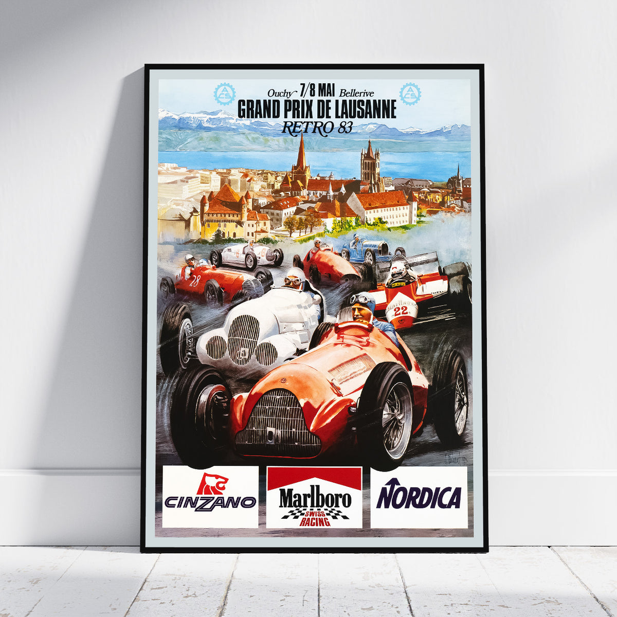 A framed vintage-style racing poster leaning against a white wall on a light floor. The poster features illustrated race cars, a cityscape, and sponsor logos including Cinzano, Marlboro, and Nordica.