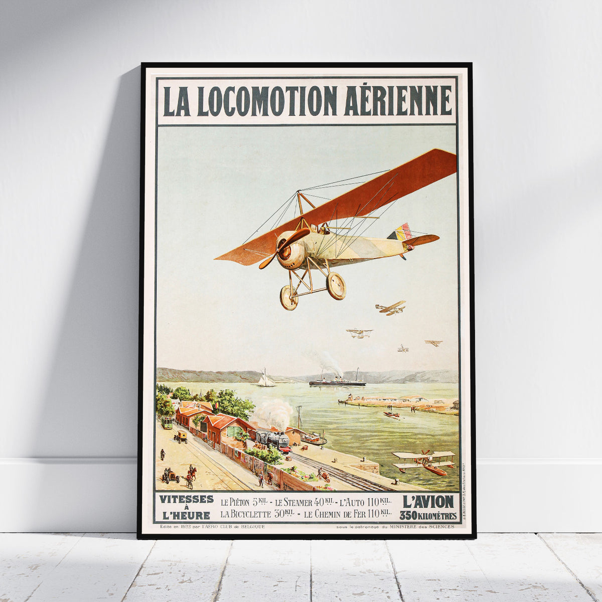 A framed vintage-style poster leaning against a white wall on a light floor. The poster reads “LA LOCOMOTION AÉRIENNE” and shows an early airplane flying over a coastal town and harbor scene.
