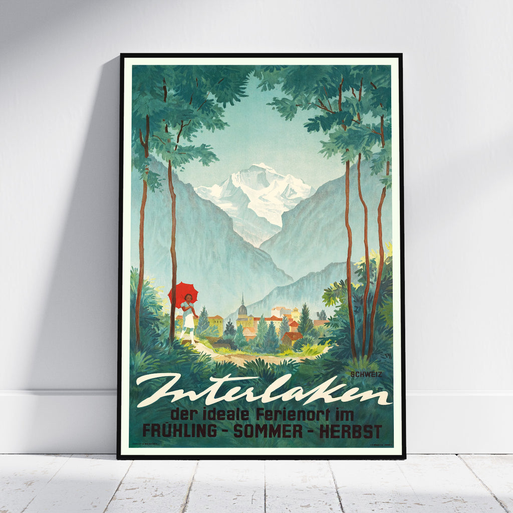 A framed vintage-style travel poster leaning against a white wall on a light wood floor. The artwork shows a scenic mountain valley with tall trees, a snow-capped peak, a small village, and a figure holding a red umbrella.