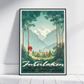 A framed vintage-style travel poster leaning against a white wall on a light wood floor. The artwork shows a scenic mountain valley with tall trees, a snow-capped peak, a small village, and a figure holding a red umbrella.