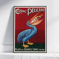 A framed vintage-style poster leaning against a white wall on a light floor. The poster shows a blue pelican holding a bottle in its beak over a red background, with the text 'Códac Pelicano' at the top and 'Hijos de Quirico López (Malaga)' at the bottom.