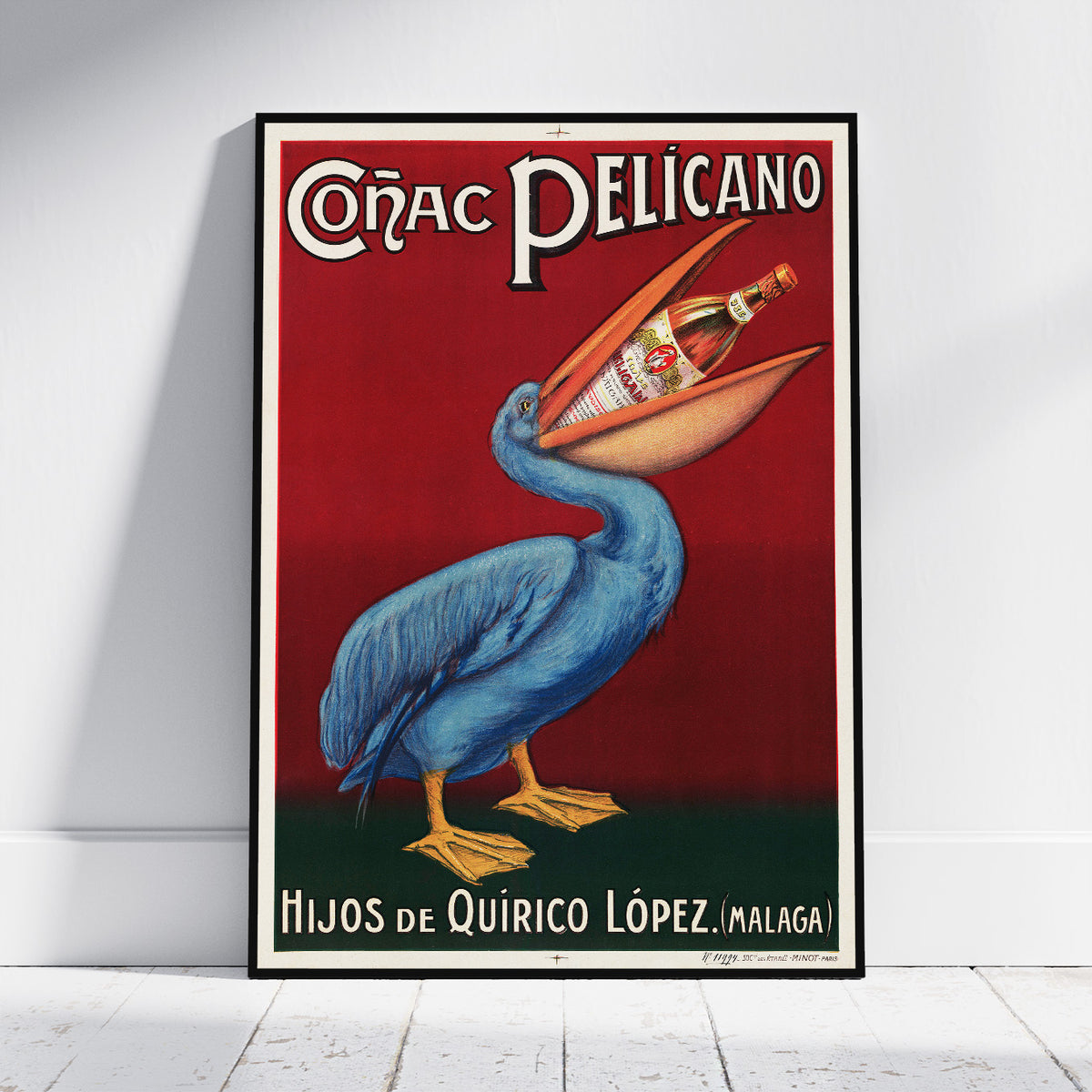 A framed vintage-style poster leaning against a white wall on a light floor. The poster shows a blue pelican holding a bottle in its beak over a red background, with the text 'Códac Pelicano' at the top and 'Hijos de Quirico López (Malaga)' at the bottom.