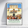 A framed vintage-style travel poster leaning against a white wall on a light wood floor. The poster features an illustration of a grand building facade with dancing figures on balconies and a large crowd gathered below, with the text 'LUGANO' and French event details.