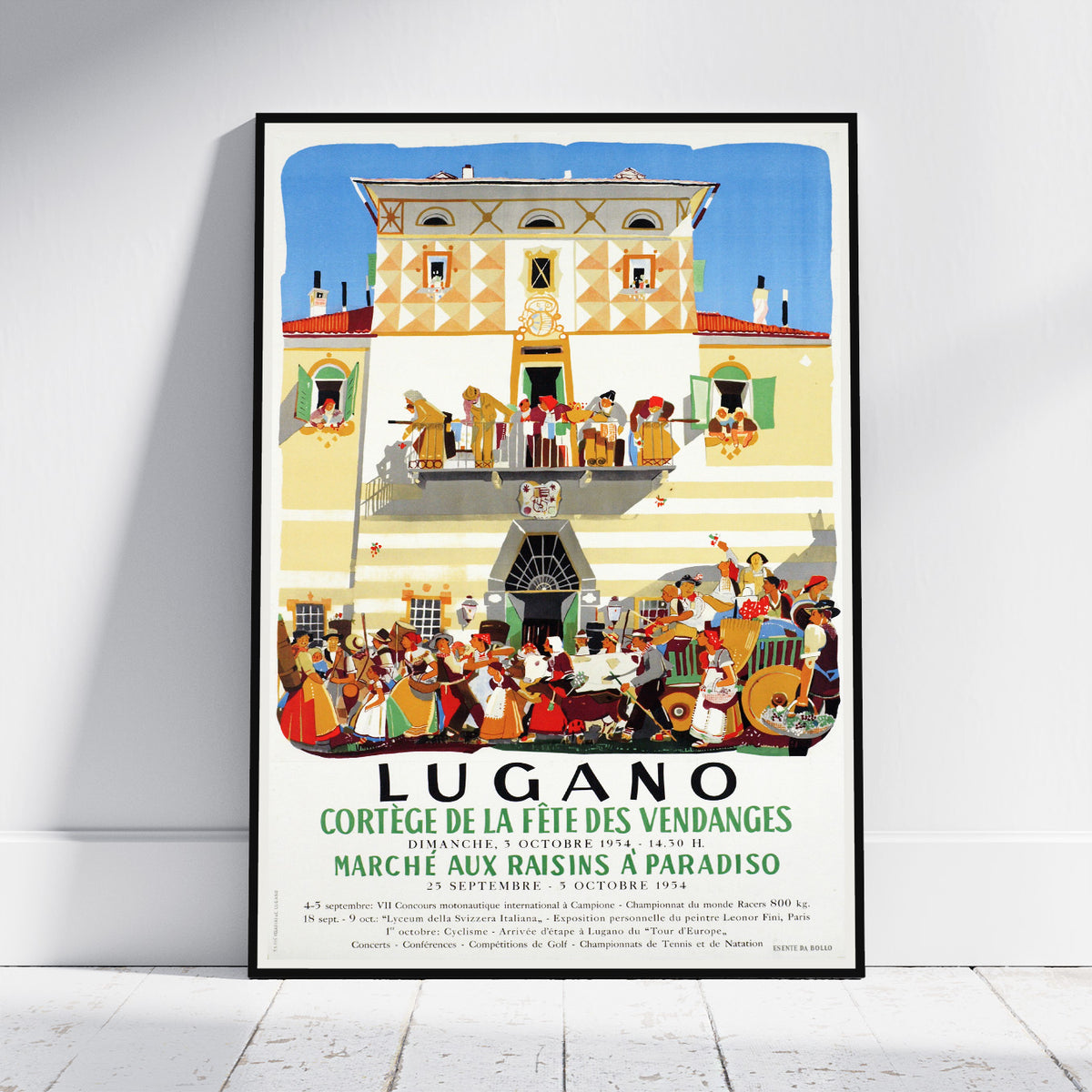 A framed vintage-style travel poster leaning against a white wall on a light wood floor. The poster features an illustration of a grand building facade with dancing figures on balconies and a large crowd gathered below, with the text 'LUGANO' and French event details.