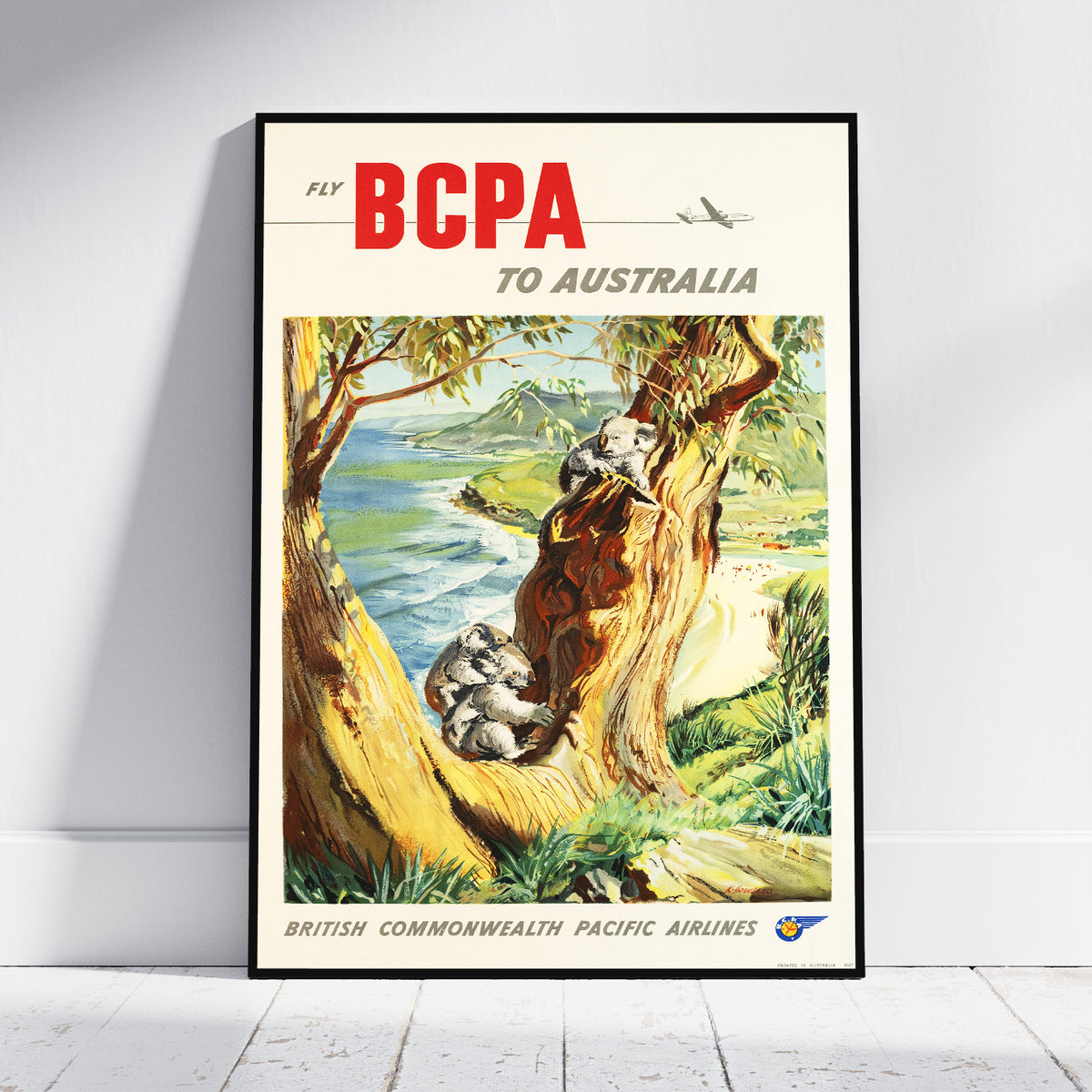 A framed vintage travel poster leaning against a white wall on a light wood floor. The poster reads 'Fly BCPA to Australia' with a colorful illustration of koalas in a tree by the sea.