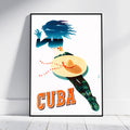 A framed travel poster of Cuba leaning against a white wall on a light wood floor. The poster features a stylized woman's silhouette filled with blue tropical waves and foliage, wearing a large yellow-orange sombrero, with the word 'CUBA' in bold orange letters at the bottom.