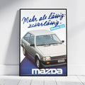 A framed vintage-style Mazda poster leaning against a white wall on a light floor. The poster features a silver Mazda 323 car with blue German text reading 'Mehr als Läufig: zuverlässig.