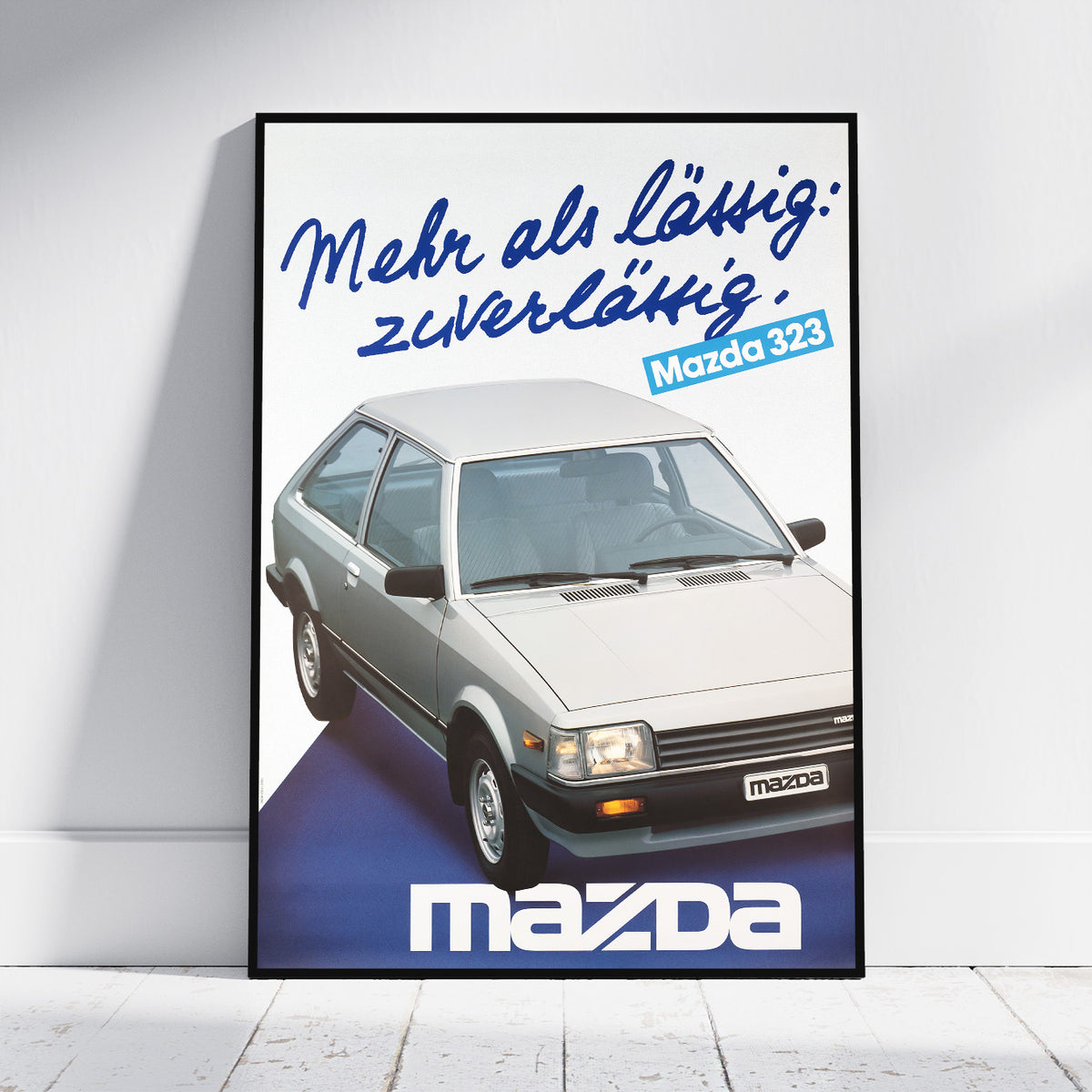A framed vintage-style Mazda poster leaning against a white wall on a light floor. The poster features a silver Mazda 323 car with blue German text reading 'Mehr als Läufig: zuverlässig.