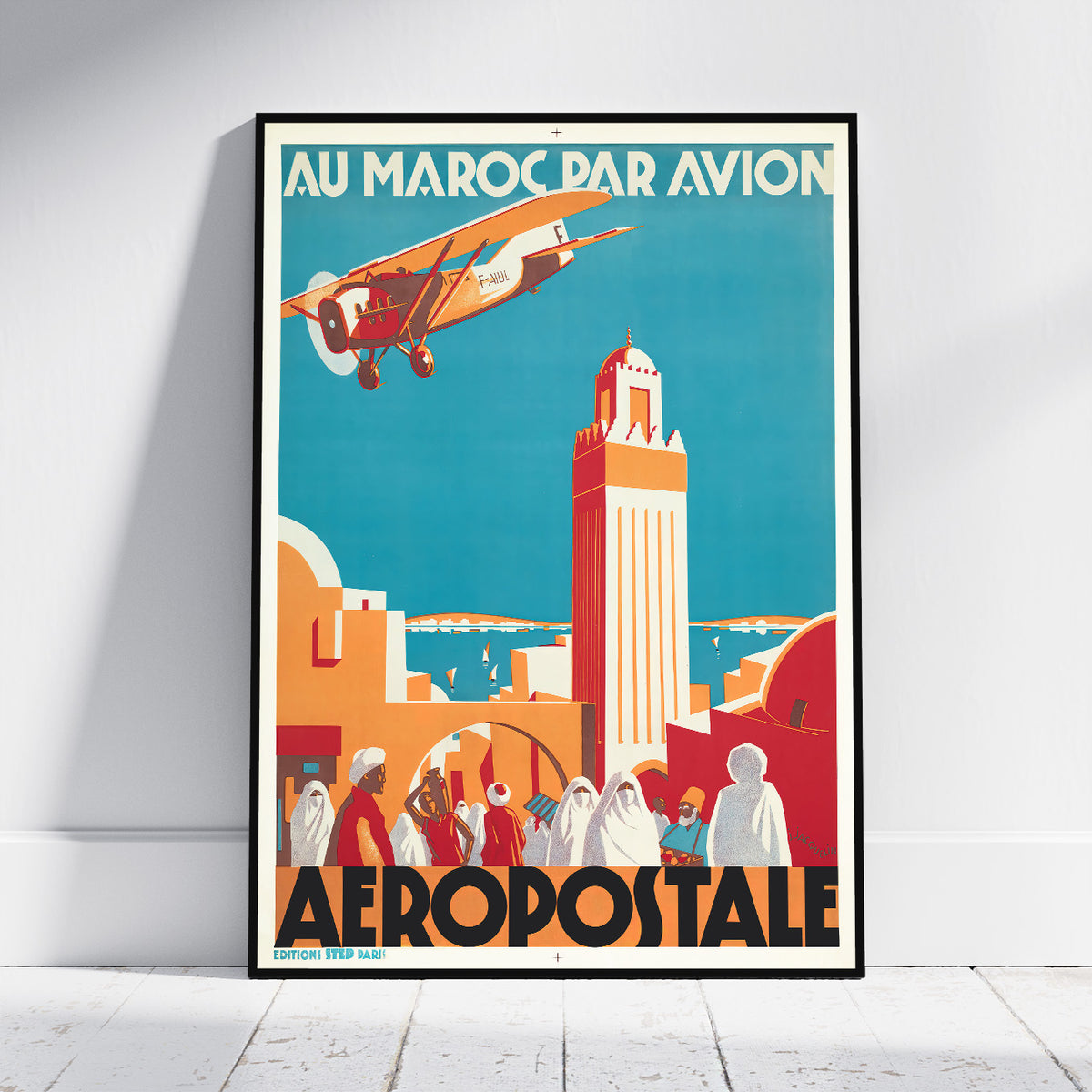 A framed vintage travel poster leaning against a white wall on a light wood floor. The poster features retro-style artwork with the text 'AU MAROC PAR AVION' at the top and 'AEROPOSTALE' at the bottom, showing an airplane over a coastal city scene in teal, orange, red, cream, and black.