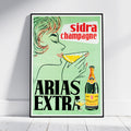 A framed vintage-style advertising poster for ARIA'S EXTRA sidra champagne leaning against a white wall on a light floor. The poster features a woman drinking from a glass, a champagne bottle, and bold text reading 'sidra champagne' and 'ARIA'S EXTRA' on a pale green background.
