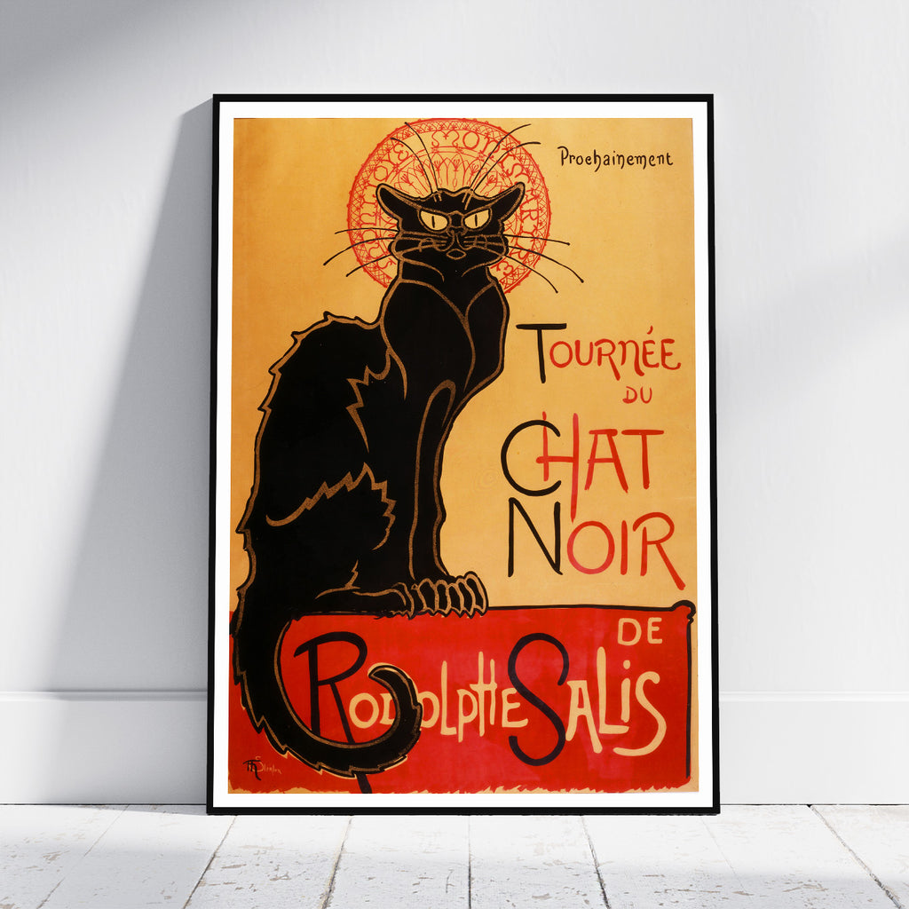 A framed vintage-style poster leaning against a white wall on a light wood floor. The poster features a black cat with a red ornamental halo and red lettering reading 'Tournée du Chat Noir de Rodolphe Salis' on a warm tan background.