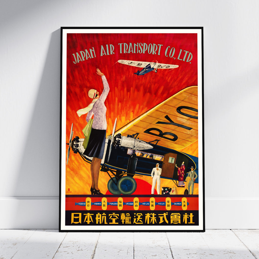 A framed vintage-style poster leaning against a white wall on a light floor. The poster features a red and orange illustrated scene with an airplane, a woman waving, and the text 'JAPAN AIR TRANSPORT CO.