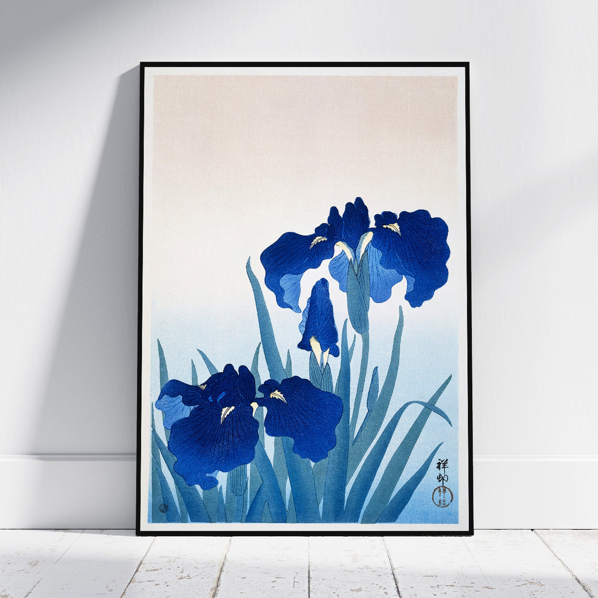 A framed art print featuring a Japanese-style illustration of deep blue irises on a soft cream and light blue background, leaning against a white wall on a light wood floor.