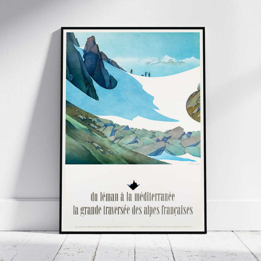A framed travel poster of a French alpine landscape print, leaning against a white wall on a light wood floor. The poster features a vintage-style illustration of snow-covered mountains, green valleys, and French text at the bottom.