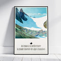 A framed travel poster of a French alpine landscape print, leaning against a white wall on a light wood floor. The poster features a vintage-style illustration of snow-covered mountains, green valleys, and French text at the bottom.