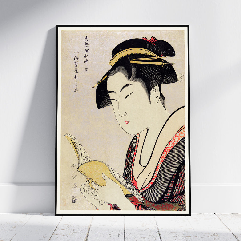 A framed Japanese ukiyo-e style art print leaning against a white wall on a light wood floor. The print depicts a woman in profile holding a book, with black hair, a cream face, and a dark kimono with red accents.