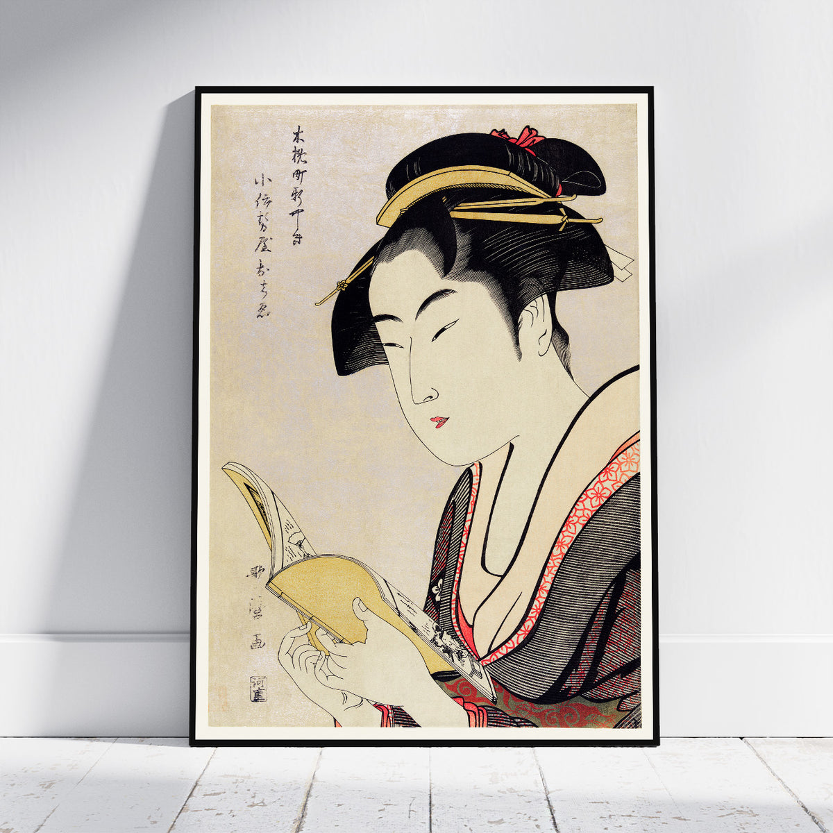 A framed Japanese ukiyo-e style art print leaning against a white wall on a light wood floor. The print depicts a woman in profile holding a book, with black hair, a cream face, and a dark kimono with red accents.