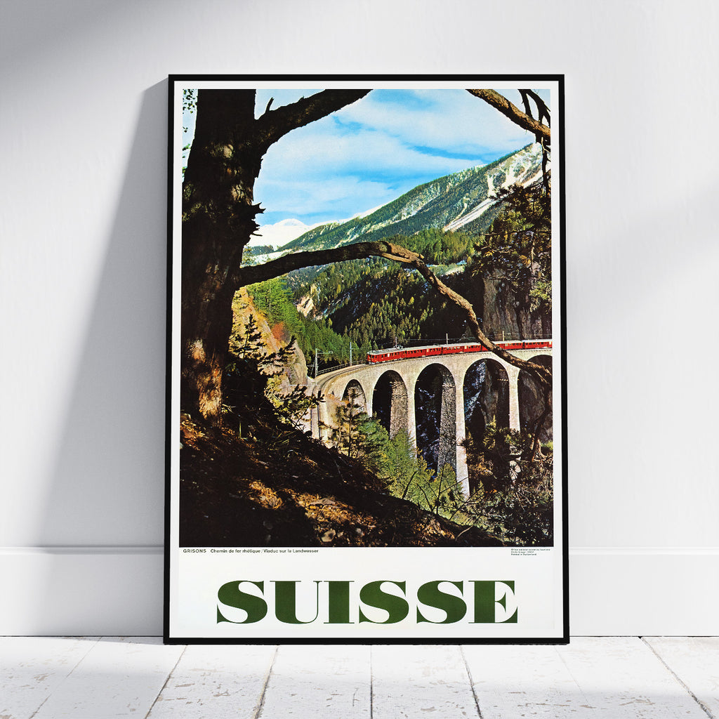 A framed vintage-style travel poster leaning against a white wall on a light wood floor. The poster depicts a scenic mountain landscape with a red train on a viaduct, large tree trunks in the foreground, and the word "SUISSE" in bold green letters at the bottom.