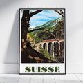 A framed vintage-style travel poster leaning against a white wall on a light wood floor. The poster depicts a scenic mountain landscape with a red train on a viaduct, large tree trunks in the foreground, and the word "SUISSE" in bold green letters at the bottom.