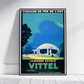 A framed vintage-style travel poster leaning against a white wall on a light wood floor. The poster features a blue-toned illustration of a domed building, trees, and bold text reading 'CHEMINS DE FER DE L'EST' and 'LA GRANDE SOURCE VITTEL'.