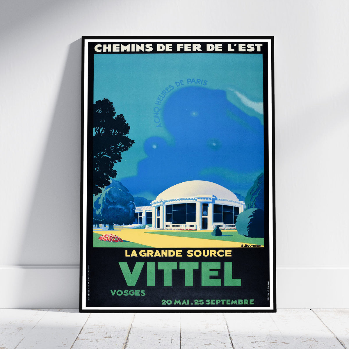 A framed vintage-style travel poster leaning against a white wall on a light wood floor. The poster features a blue-toned illustration of a domed building, trees, and bold text reading 'CHEMINS DE FER DE L'EST' and 'LA GRANDE SOURCE VITTEL'.