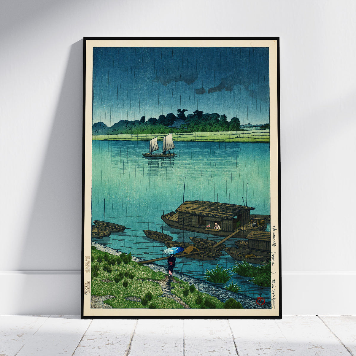 A framed Japanese-style art print leaning against a white wall on a light wood floor. The artwork depicts a rainy river scene with a sailboat, houseboats, green shoreline, and a person holding an umbrella near the shore.