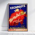 A framed vintage French advertising poster for 'Ménetre et Cie' hangs against a white wall on a light wood floor. The poster features a deep blue background, a large red leaf, a cartoon snail, and bold white and yellow text reading 'ESCARGOTS', 'Ménetre', 'EXQUIS EXQUIS!', and 'Aux Parcs de Bourgogne'.