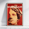 A framed poster leaning against a white wall on a light floor. The poster features a stylized classical bust portrait in warm beige tones over a vivid red background, with the word 'SWISSAIR' in large white letters at the top.