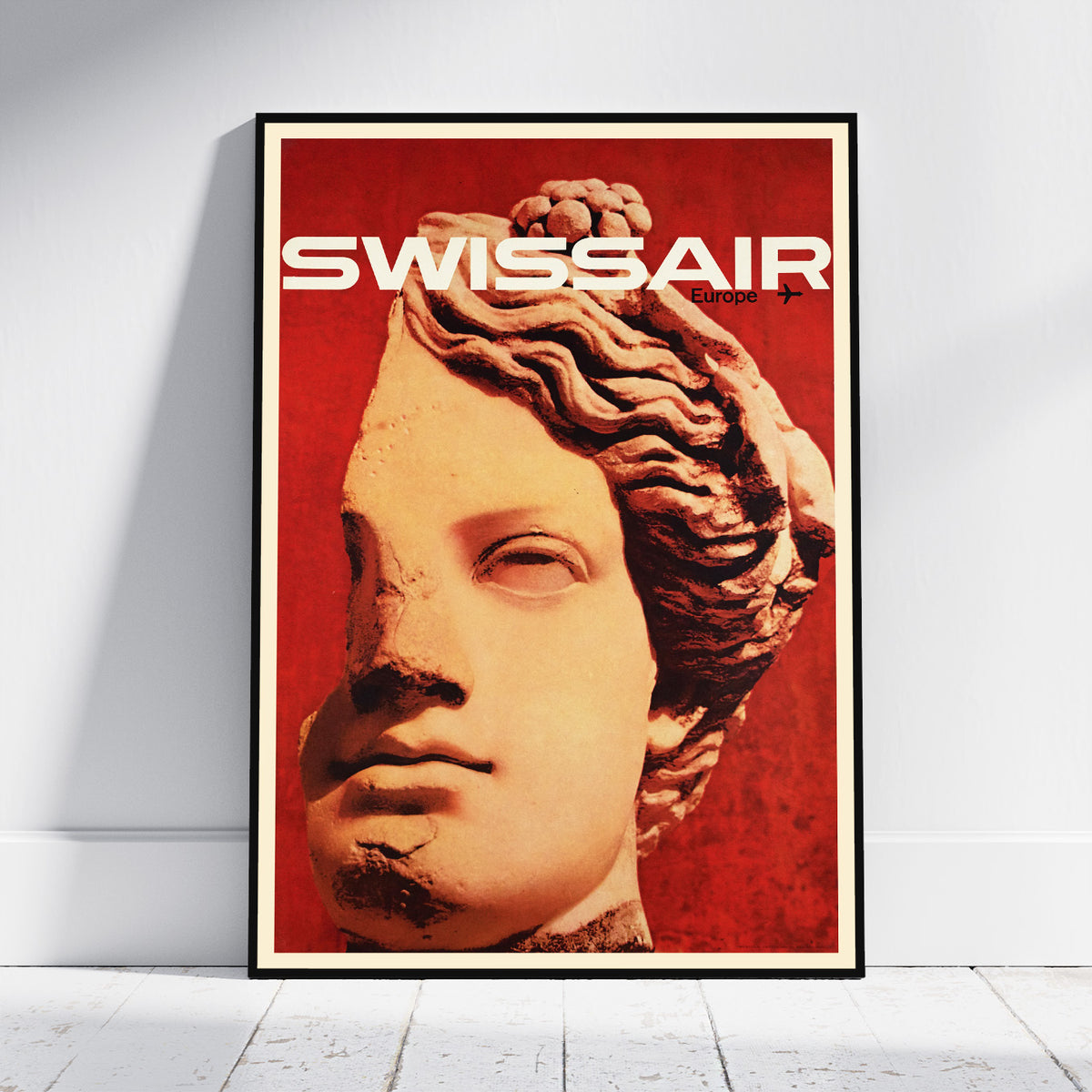 A framed poster leaning against a white wall on a light floor. The poster features a stylized classical bust portrait in warm beige tones over a vivid red background, with the word 'SWISSAIR' in large white letters at the top.